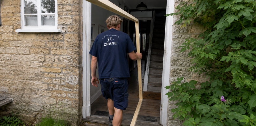 A Crane fitter carries the front end of a rectangular test frame through the front door of a cottage